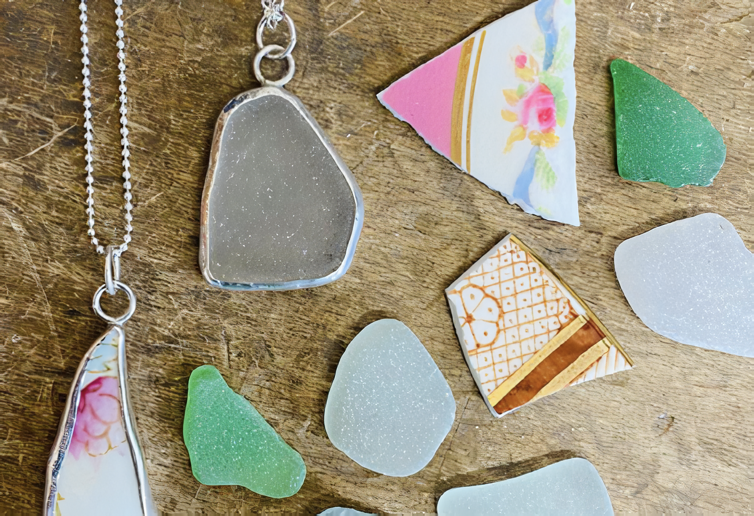 Two silver chain necklaces with resin and metal pendants surrounded by an assortment of sea glass on a wooden surface.